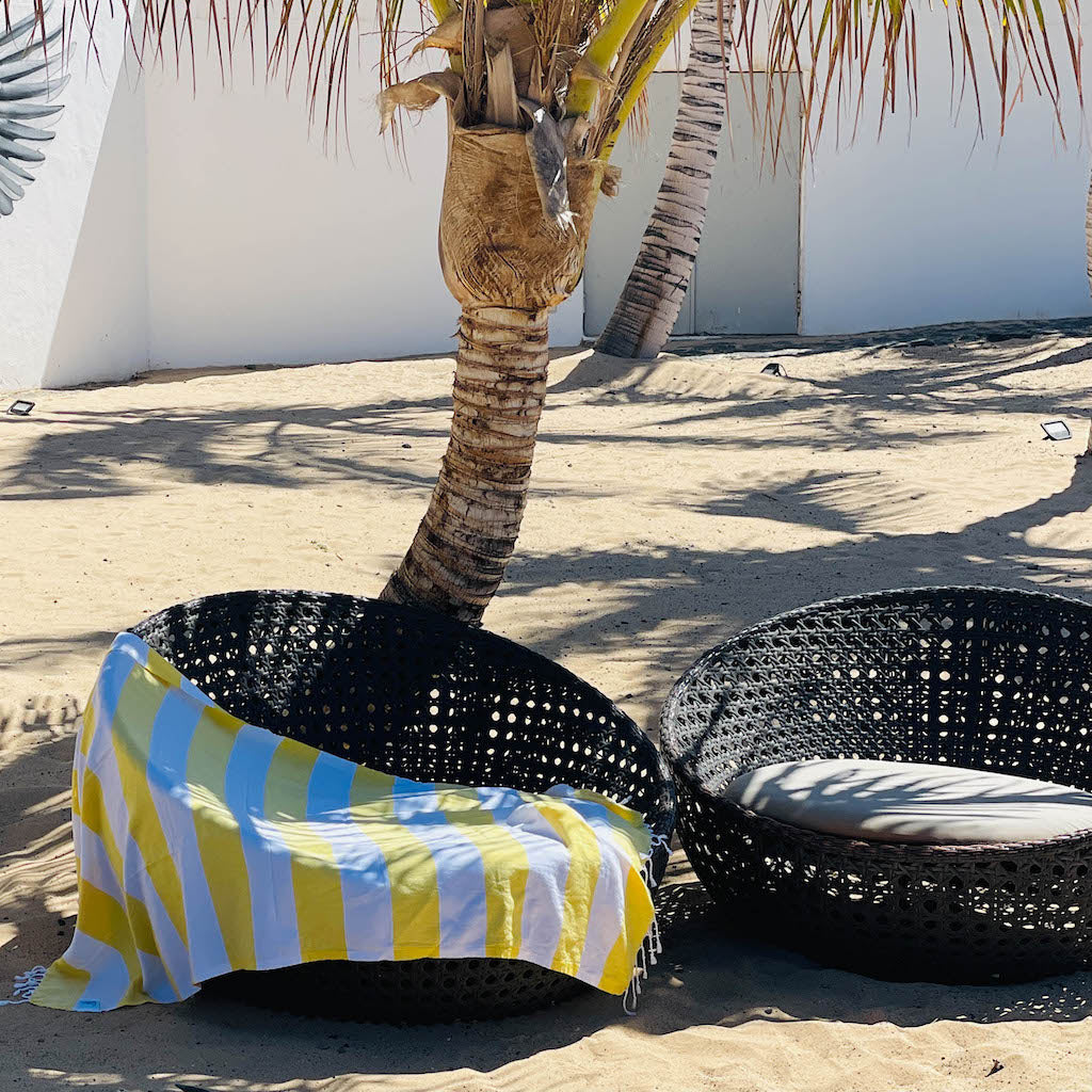 Black wicker chair with a yellow and blue striped towel on a sandy beach with palm trees.