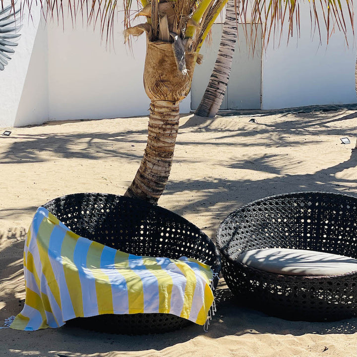 Black wicker chair with a yellow and blue striped towel on a sandy beach with palm trees.